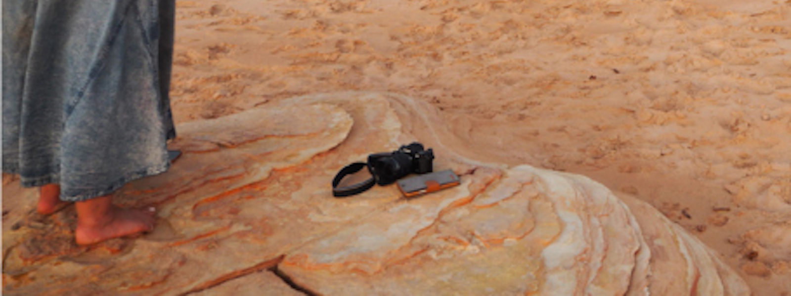 A photographer stands barefoot on sandy ground, taking a photo of three women standing together in front of a striking red cliff face. The women are smiling and wearing patterned dresses, framed by the warm tones of the landscape, suggesting a relaxed, collaborative moment outdoors.